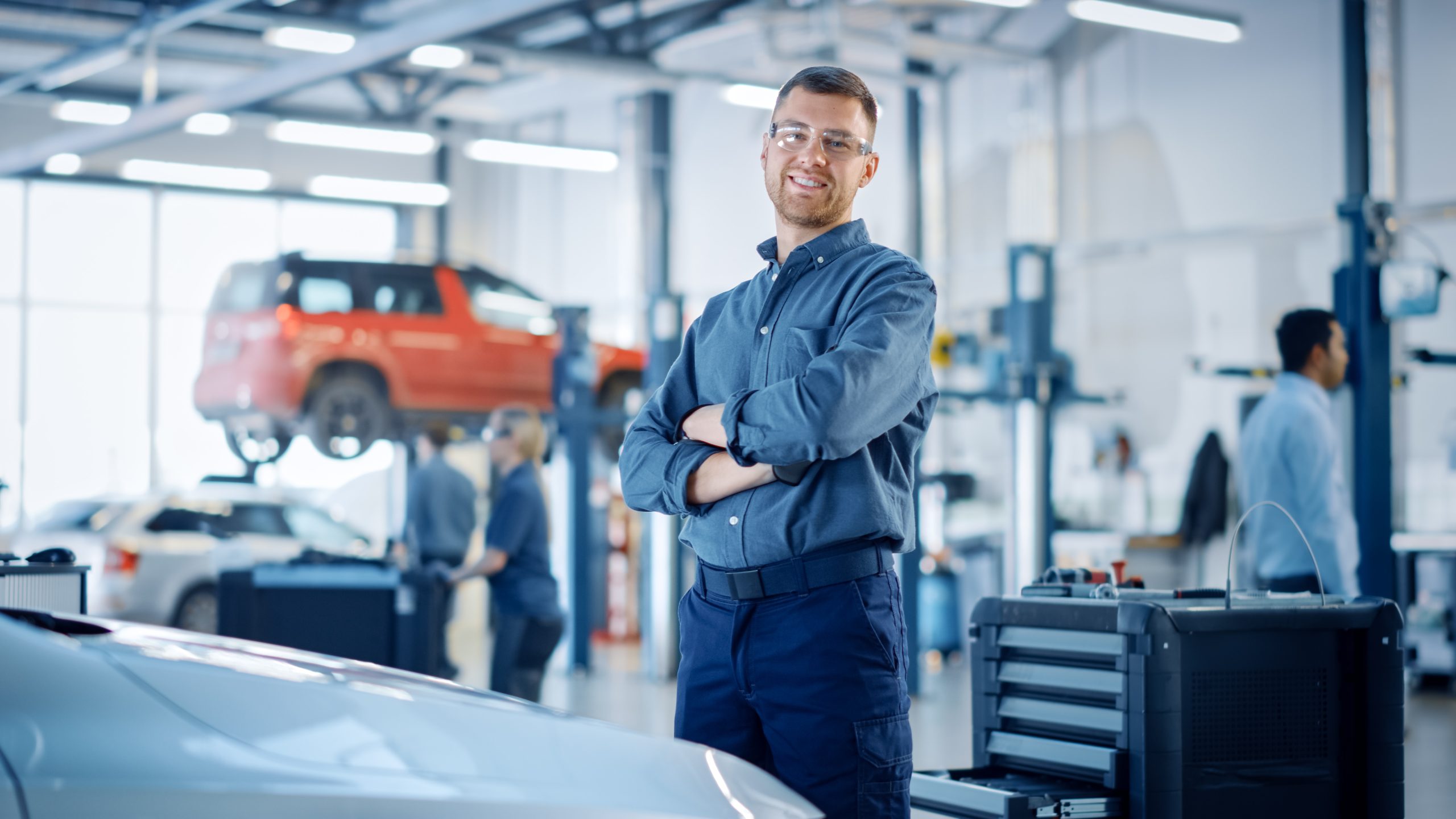 A photo of a mechanic in a blue uniform standing in a repair shop with his arms crossed.